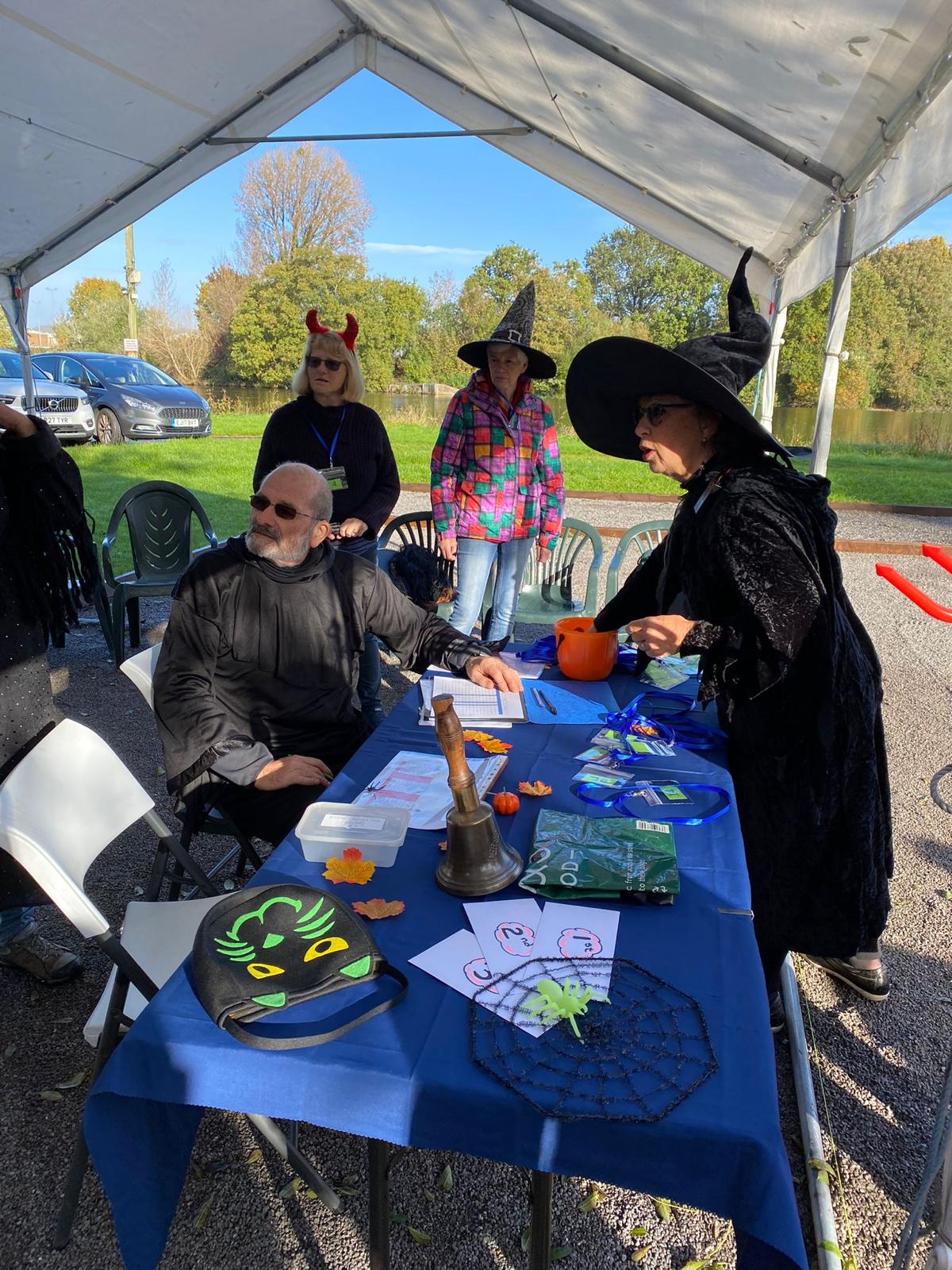 Members in Halloween costumes, including witches’ hats and devil horns, at the mêlée registration table under the gazebo