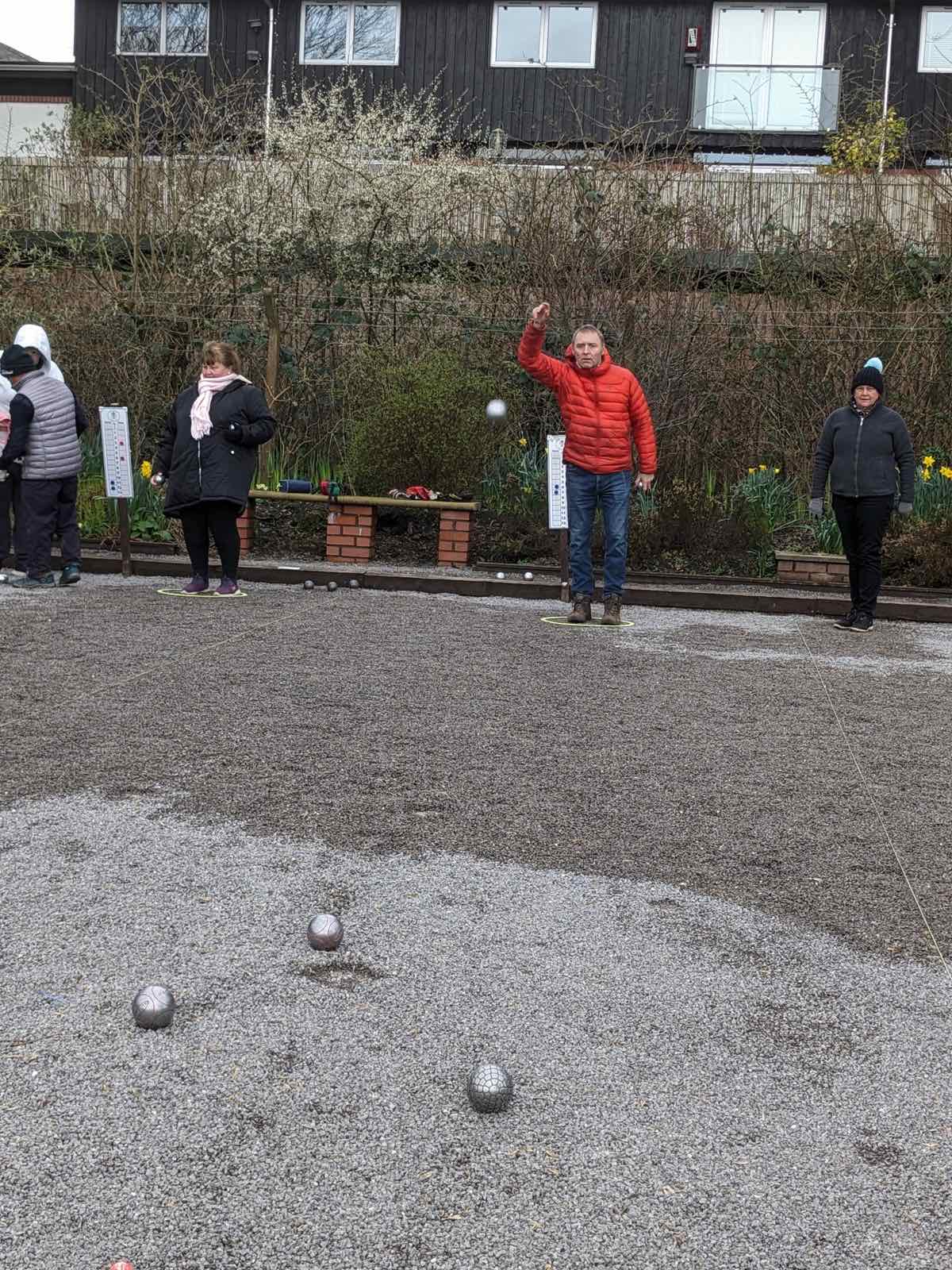 A club member in a red jacket throwing a boule on the piste while other players look on